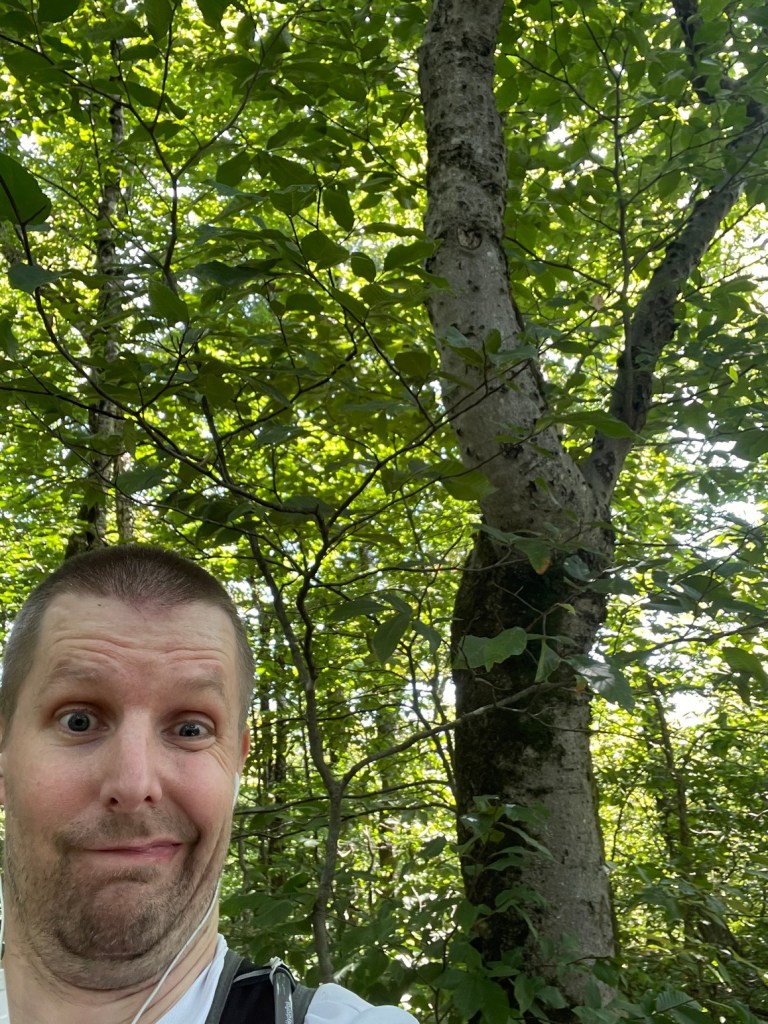 A person making a funny facial expression in a forested area with green leaves and trees in the background.
