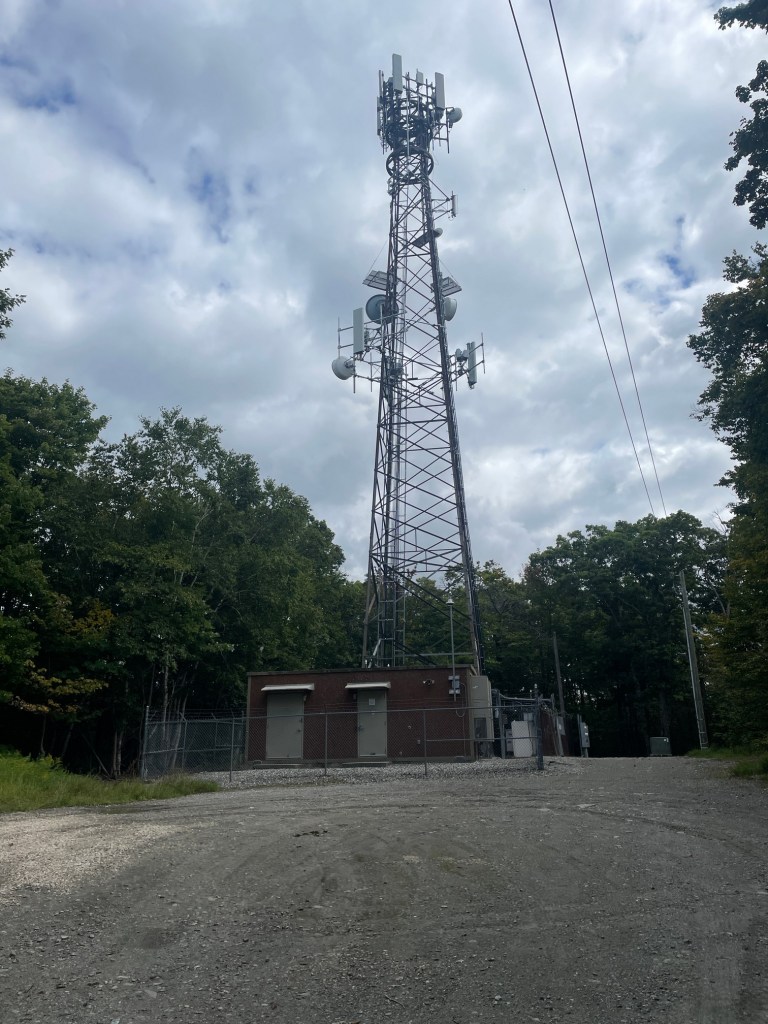 A tall telecommunications tower with antennas, situated near a brick building, surrounded by trees and a gravel path.